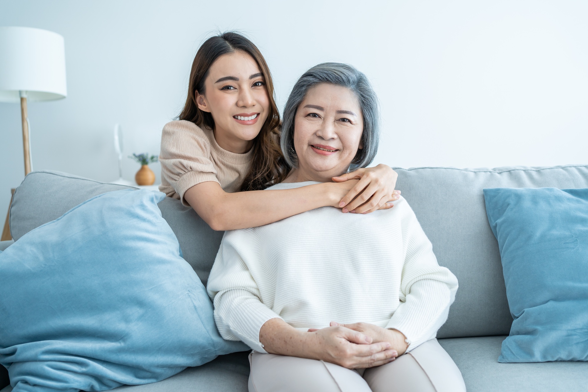 Attractive woman and senior elder mom sit on sofa together in living room and smile, look at camera