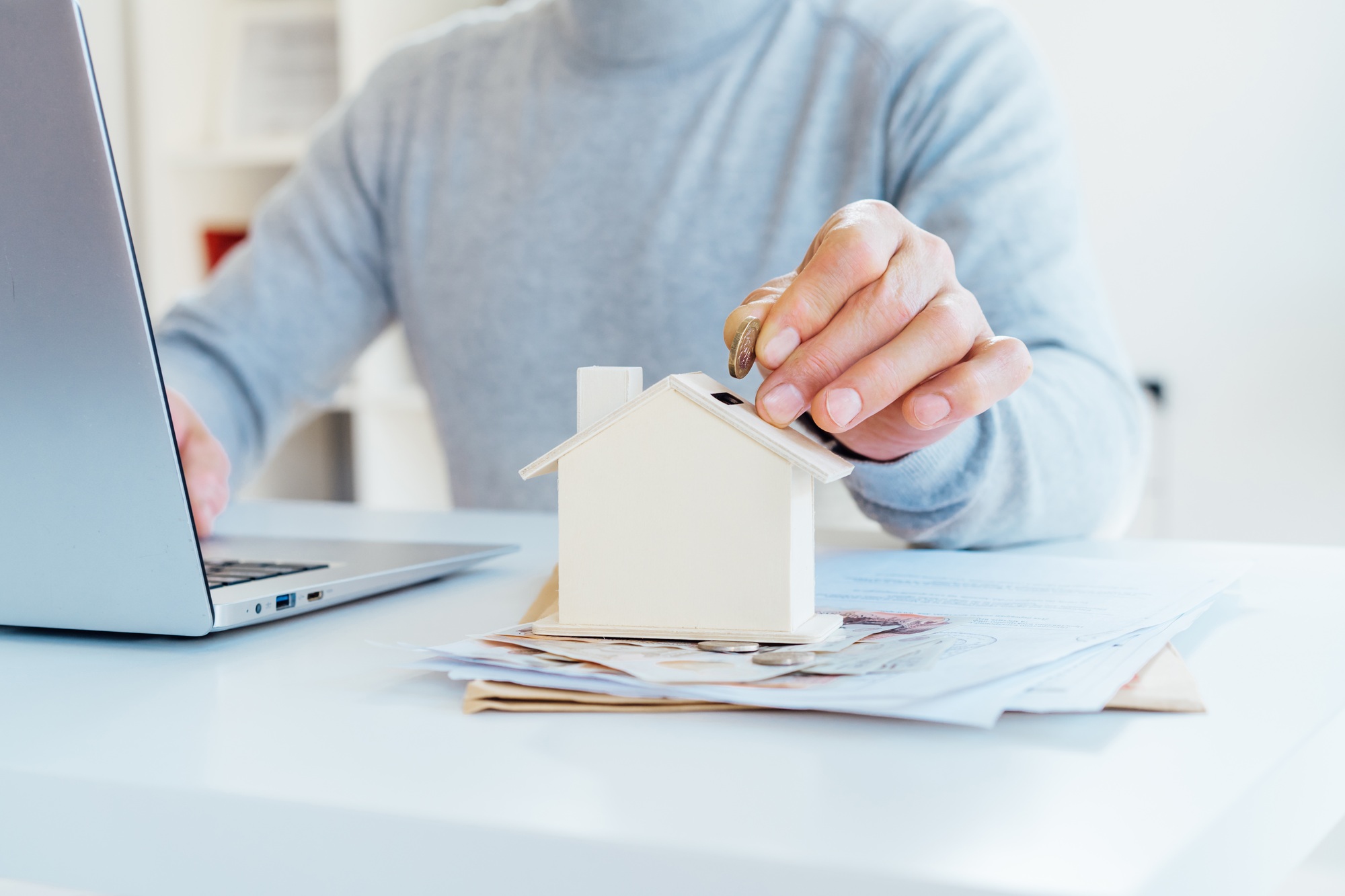 Close up male hand putting coin into House model money box on table with money and documents.